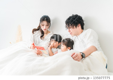 ベッドで子供に絵本を読んであげる母親と父親(寝かしつけ,お昼寝,読み聞かせ) ベッドで子供に絵本を読んであげる母親と父親(寝かしつけ,お昼寝,読み聞かせ) 135077775