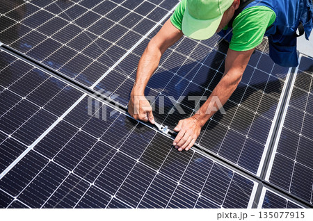 Worker building photovoltaic solar panel system on rooftop of house. Close up of man engineer installing solar module with help of wrench outdoors. Alternative and renewable energy generation concept. 135077915
