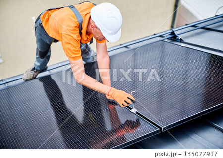 Worker building photovoltaic solar panel system on rooftop of house. Man engineer in helmets and gloves installing solar module with help of hex key outdoors. Alternative energy generation concept Worker building photovoltaic solar panel system on rooftop of house. Man engineer in helmets and gloves installing solar module with help of hex key outdoors. Alternative energy generation concept 135077917
