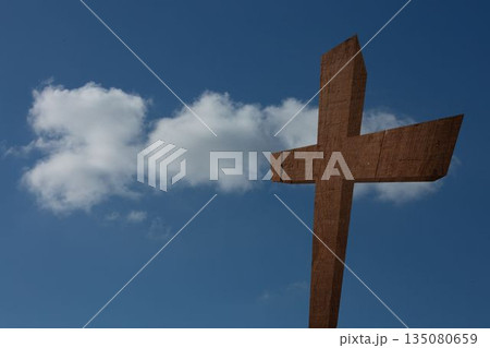 Rough-hewn wooden cross is standing against clear blue sky with drifting white cloud in flat design 135080659