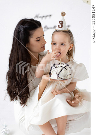 A mother and her daughter celebrate the little girl's third birthday with a cake and a party hat. A mother and her daughter celebrate the little girl's third birthday with a cake and a party hat. 135081147