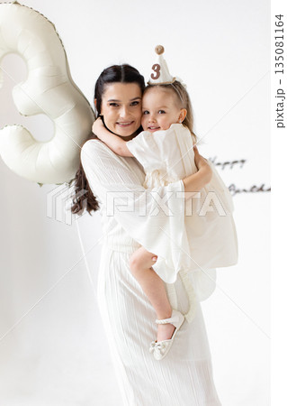 A mother embraces her daughter, celebrating her third birthday with a balloon and a party hat. 135081164