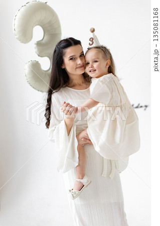 A mother holds her daughter, celebrating her third birthday with a balloon and party hat. A mother holds her daughter, celebrating her third birthday with a balloon and party hat. 135081168