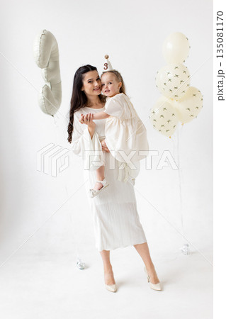 A mother holds her daughter, celebrating her third birthday with balloons and a party hat, all in white. A mother holds her daughter, celebrating her third birthday with balloons and a party hat, all in white. 135081170
