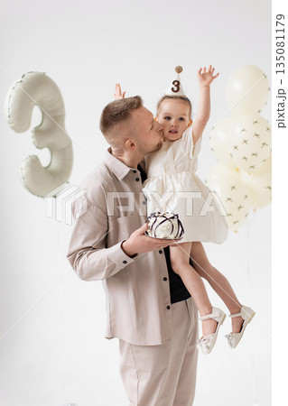 A father kisses his daughter on the cheek while holding her at her third birthday celebration, complete with a cake and balloons. A father kisses his daughter on the cheek while holding her at her third birthday celebration, complete with a cake and balloons. 135081179