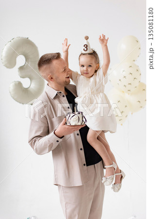 A father holds his daughter, celebrating her third birthday with a cake and balloons. A father holds his daughter, celebrating her third birthday with a cake and balloons. 135081180