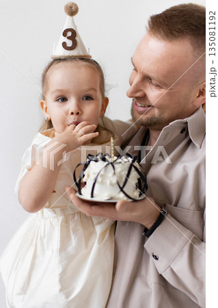 A young girl celebrates her third birthday with her father, holding a cake with a candle. 135081192