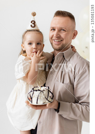 A father holds his daughter, who is celebrating her third birthday with a cake. 135081193