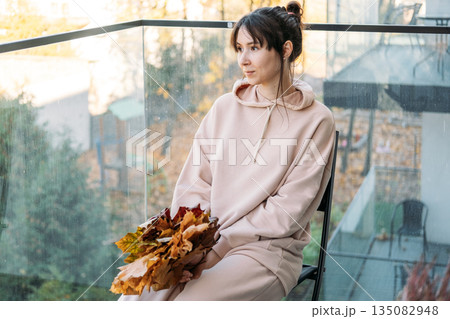 Woman looks out from balcony while holding bouquet of autumn leaves. Slow living, conscious consumption, sustainability, seasonal living. 135082948