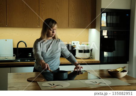 Woman stands in modern kitchen with sleek appliances while preparing meal on induction stove. Contemporary kitchen design, functional living, minimalist home, sleek appliances. 135082951
