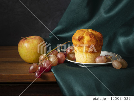 Still Life With Apple, Grapes, And Muffin On Plate Beside Dark Green Fabric Backdrop 135084525