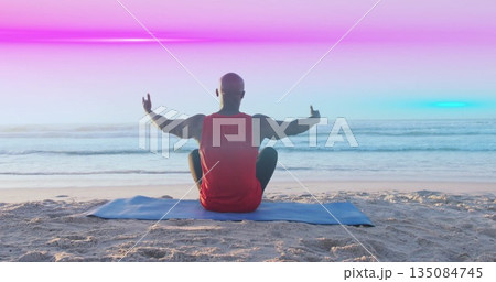 Sitting man wearing red tank top, shorts, raising thumbs on sandy beach, with blue exercise mat 135084745