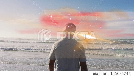 Senior man in casual shirt gazing at ocean at beach, with lens flare and cosmic sparkles 135084746