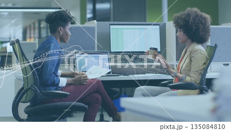 Businesswomen discussing report at desk in modern office, with laptop, dual monitors, coffee cup 135084810