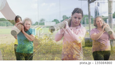 Striding women across grassy field carrying logs on shoulders, with metal rig and chain-link fence 135084860