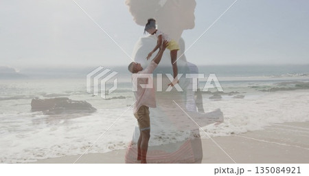 Man in pink shirt lifting girl in yellow shorts at beach water's edge, with mother silhouette 135084921