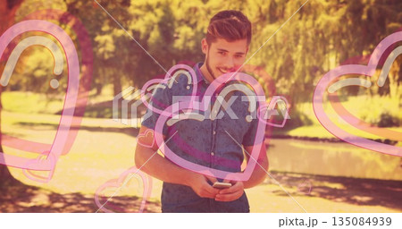 Man wearing denim reading texts on smartphone in park beside pond, with pink heart overlays Man wearing denim reading texts on smartphone in park beside pond, with pink heart overlays 135084939