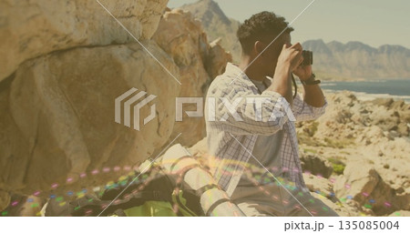 Hiker using binoculars and sitting on rocky ledge by sea, with backpack and rolled sleeping mat Hiker using binoculars and sitting on rocky ledge by sea, with backpack and rolled sleeping mat 135085004