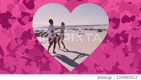 Spinning couple holding hands and dancing on sandy beach, with ocean waves and heart frame motifs 135085012
