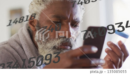 Holding smartphone and pill bottle, senior man in bathrobe viewing numeric data overlays at home 135085055