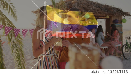 Woman in striped sundress and man in polo shirt chatting at beach bar with drinks, bunting 135085118