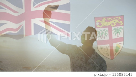 Holding smartphone, man in floral shirt taking selfie on shore, with Fiji flag, coat arms overlays 135085143