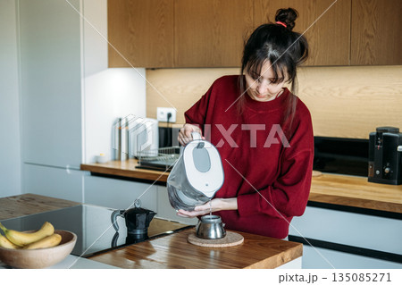 Person pours filtered water from pitcher into base of moka pot on wooden counter. Manual espresso routine, elevated home brewing, artisanal coffee gear, precision coffee making. Person pours filtered water from pitcher into base of moka pot on wooden counter. Manual espresso routine, elevated home brewing, artisanal coffee gear, precision coffee making. 135085271