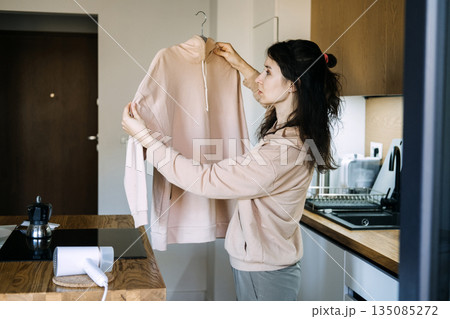 Woman examines hoodie sleeve while holding garment on hanger in kitchen. Reducing fashion waste, fabric preservation, gentle steaming. Woman examines hoodie sleeve while holding garment on hanger in kitchen. Reducing fashion waste, fabric preservation, gentle steaming. 135085272