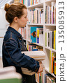 woman holds stack of books among shelves in library. student is getting education. 135085913