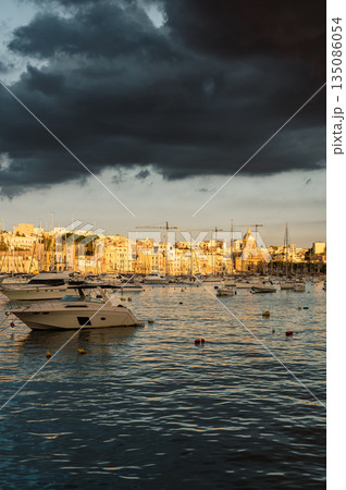 A picturesque view of Birgu Port filled with numerous boats, bathed in warm pre-sunset light. The tranquil waters reflect the golden hues, creating a serene maritime scene in Malta. 135086054