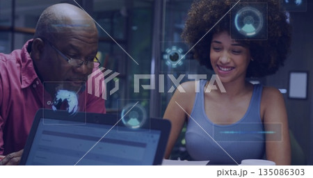 Collaborating man and woman reviewing documents in office with laptop, digital interface graphics 135086303