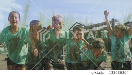Laughing muddy children in matching green shirts leaning on muddy field, with wooden obstacle poles 135086364