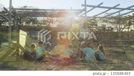 Teaching instructor holding paper and speaking to children under vine pergola, with campfire ring 135086365