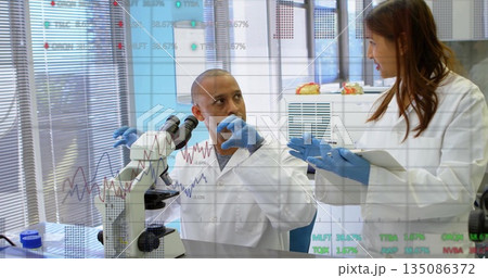Two researchers discussing findings at lab bench, with microscope, tablet, gloves and sample trays Two researchers discussing findings at lab bench, with microscope, tablet, gloves and sample trays 135086372