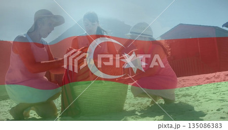 Kneeling grandmother mother daughter unfolding striped towel on sandy beach, wearing sunhats 135086383