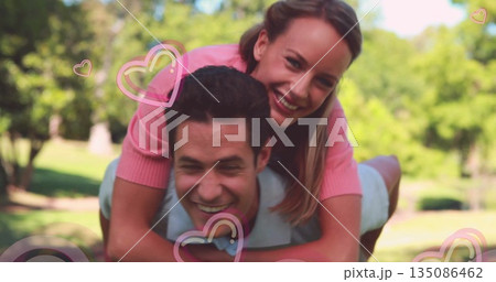 Carrying couple wearing grey polo and pink top posing in sunlit park, with pink heart graphics 135086462