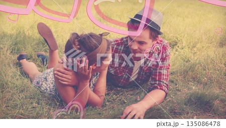 Lying couple wearing red plaid shirt, floral romper sharing gazes on meadow with pink heart doodles 135086478
