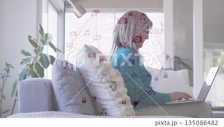 Typing woman wearing teal sweater leaning on sofa in living room, with laptop and network icons 135086482