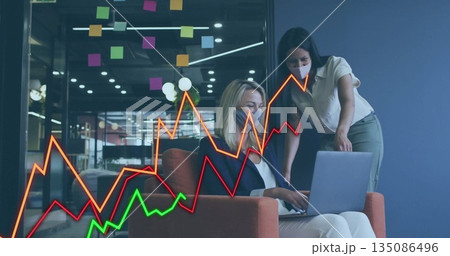 Collaborating women wearing face masks reviewing laptop line graph in lounge, with sticky notes 135086496