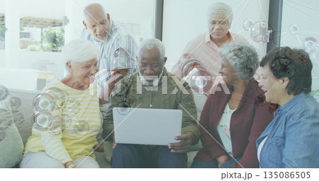 Gathering seniors wearing casual clothing collaborating around silver laptop in residential lounge Gathering seniors wearing casual clothing collaborating around silver laptop in residential lounge 135086505