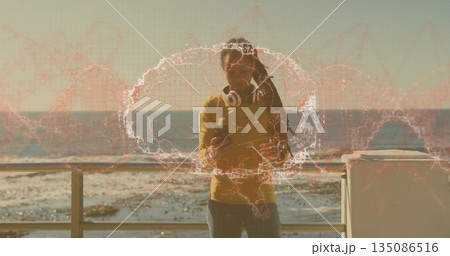 Leaning on railing, woman wearing mustard shirt holding smartphone at promenade, wearing headphones 135086516