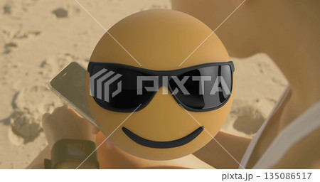 Round yellow emoticon with sunglasses floating over woman on beach, with smartphone, smartwatch 135086517