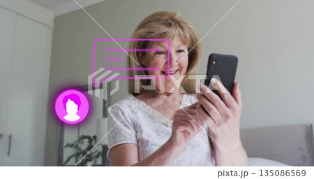 Smiling senior woman using smartphone on bed in bedroom, with lamp, plant, mirror and chat icons Smiling senior woman using smartphone on bed in bedroom, with lamp, plant, mirror and chat icons 135086569
