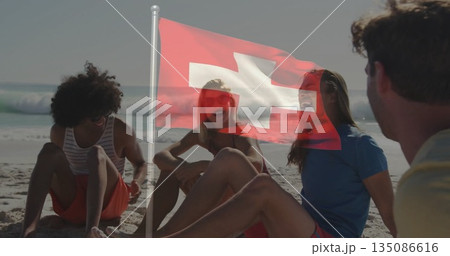 Sitting friends chatting in swimwear on sandy beach near shoreline, with Swiss flag and sunglasses 135086616
