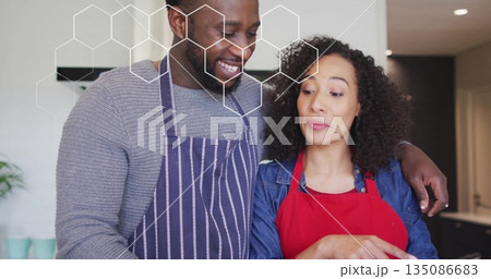 Couple hugging and cooking in home kitchen, with striped apron, red apron and hexagonal overlay 135086683