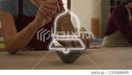 Potter wearing burgundy apron shaping clay vessel in pottery studio, with wooden tool and slip bowl Potter wearing burgundy apron shaping clay vessel in pottery studio, with wooden tool and slip bowl 135086690