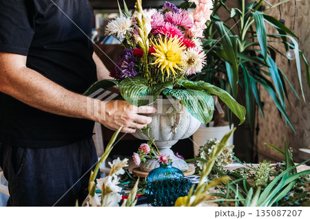 Hands hold finished floral arrangement in stone urn featuring colorful asters and large green hosta leaves. Floral design class, DIY flower arranging, floristry workshop. Hands hold finished floral arrangement in stone urn featuring colorful asters and large green hosta leaves. Floral design class, DIY flower arranging, floristry workshop. 135087207
