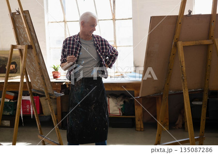 Wearing paint stained apron, senior male painter holding paintbrush in studio between two easels Wearing paint stained apron, senior male painter holding paintbrush in studio between two easels 135087286