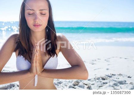 Woman in 20s meditating on sandy beach wearing white bikini top and pressing hands together Woman in 20s meditating on sandy beach wearing white bikini top and pressing hands together 135087300