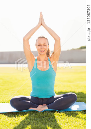 Woman practicing yoga cross-legged on yoga mat on green lawn wearing turquoise tank top, leggings Woman practicing yoga cross-legged on yoga mat on green lawn wearing turquoise tank top, leggings 135087303